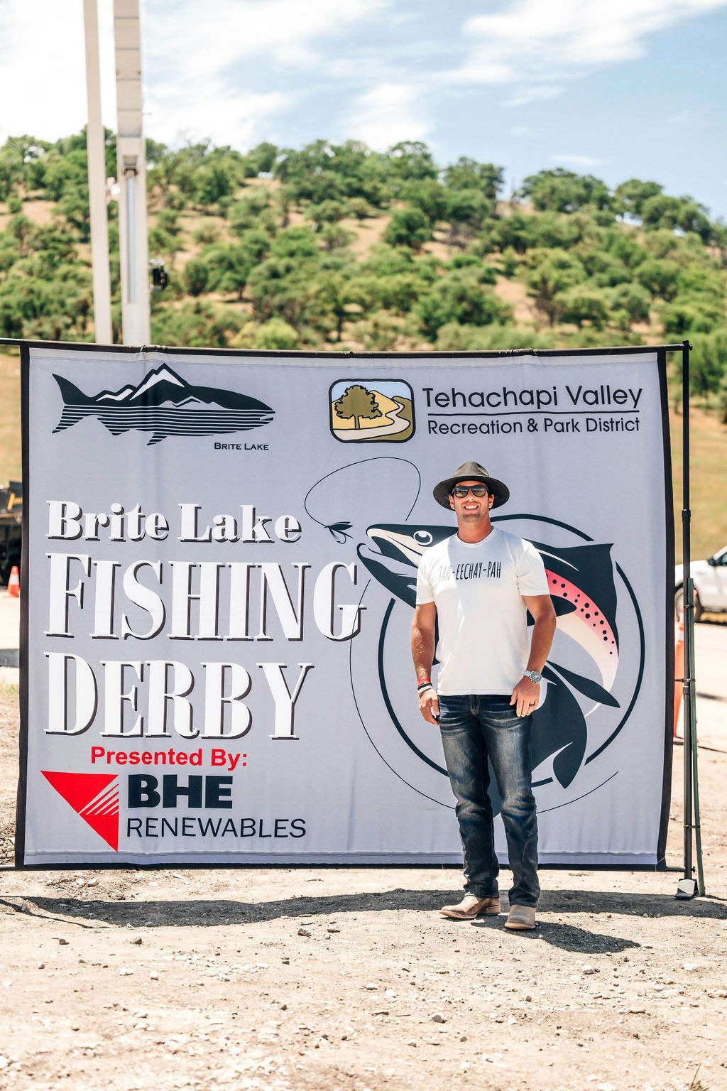 A man stands in front of a Brite Lake Fishing Derby banner, outdoors on a sunny day.