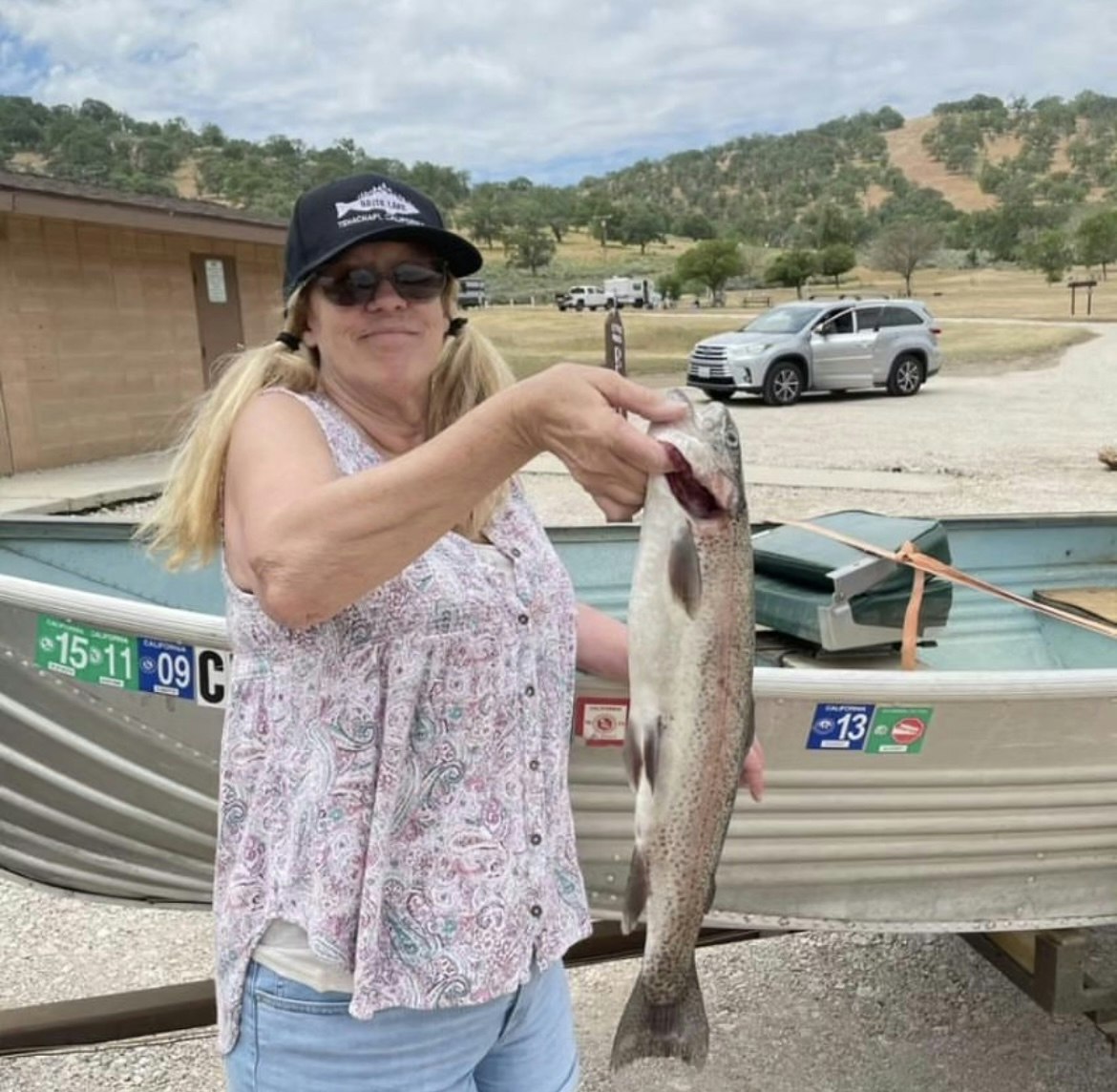 A person holding a large fish by a boat, with vehicles and hills in the background.