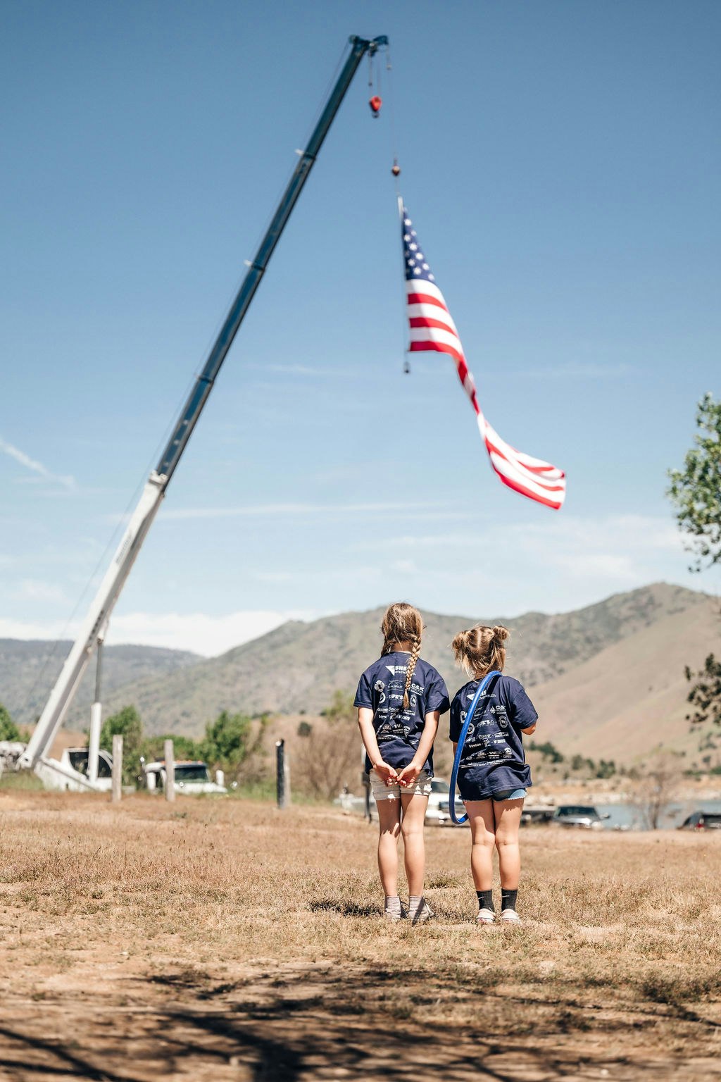 Two people looking at a large American flag hanging from a crane against a mountainous backdrop.