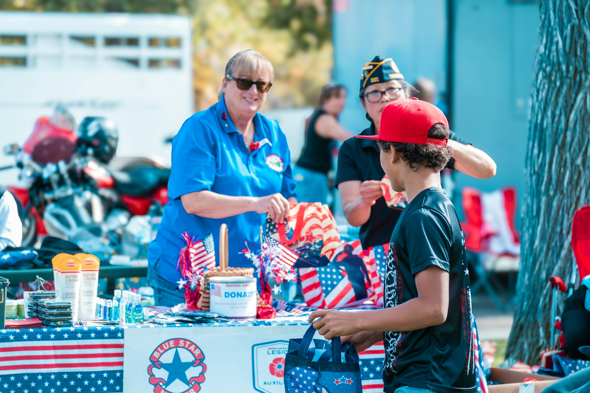 People at a booth with American flags, donation signs, and festive items; a child holding a patriotic bag.