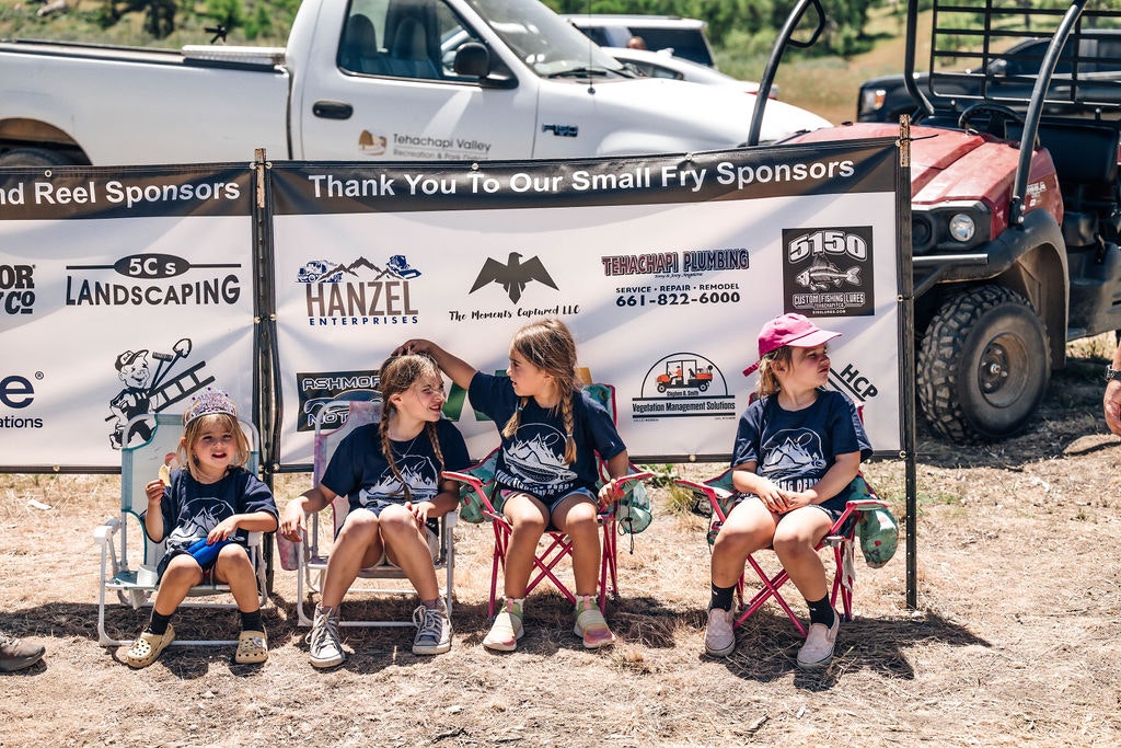 Three children are sitting on folding chairs in front of sponsor banners outdoors, with vehicles and greenery in the background.