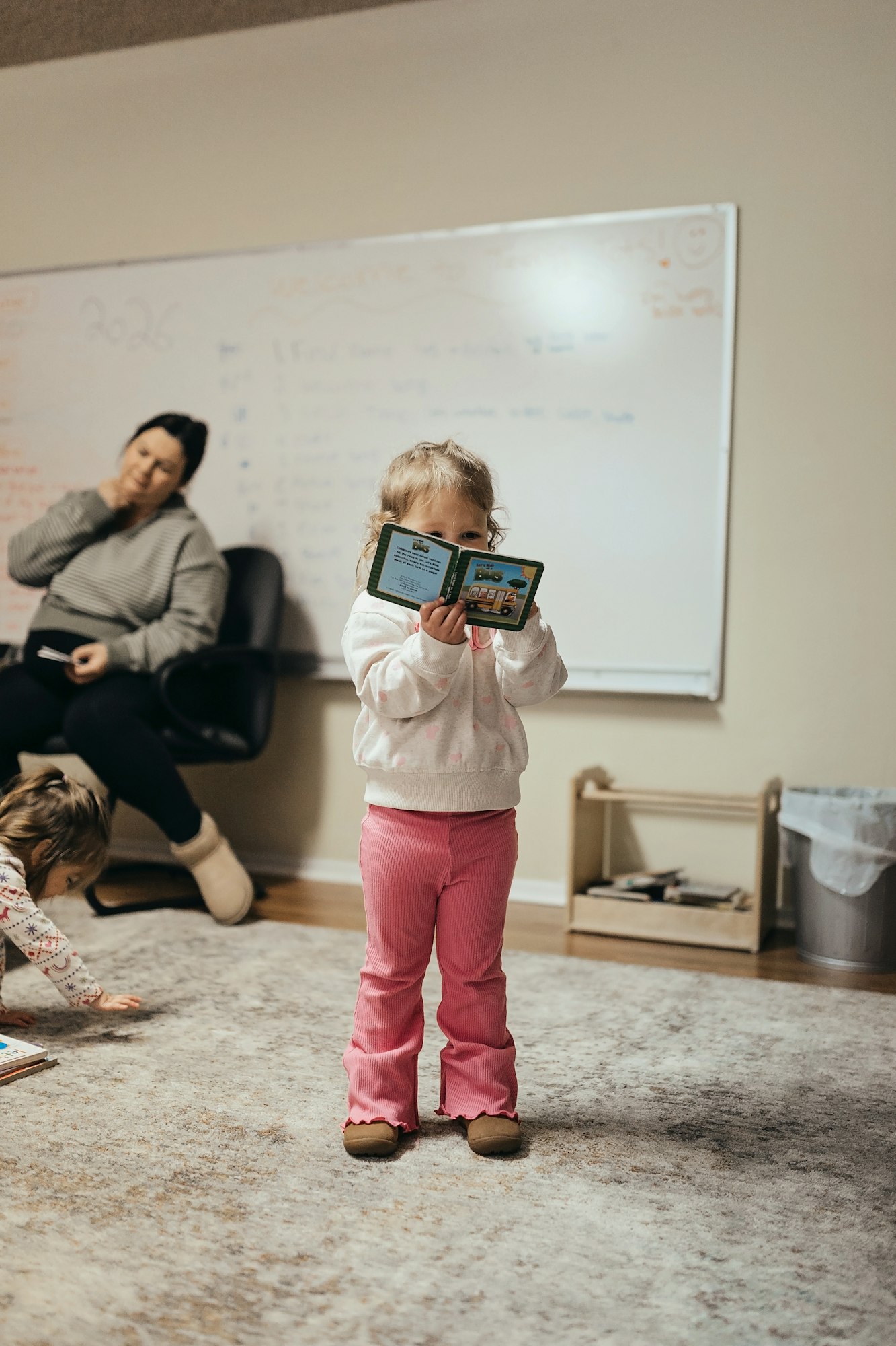 A young girl in pink pants holds a book, while a woman in the background appears to be deep in thought.