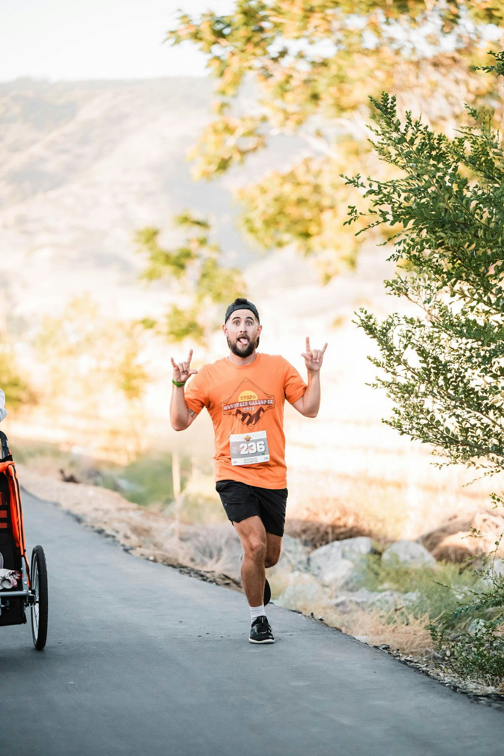 A man running a marathon in an orange shirt with a race bib, making a playful gesture, surrounded by trees.