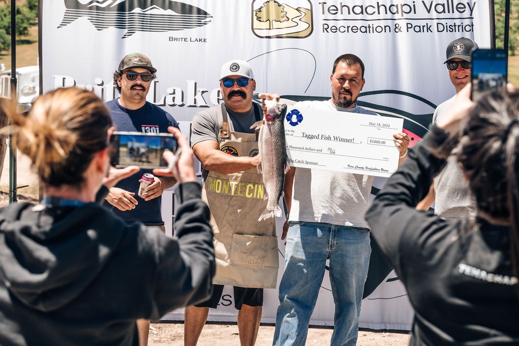 A person holding a check and a fish, celebrating a win, with onlookers capturing the moment on camera.