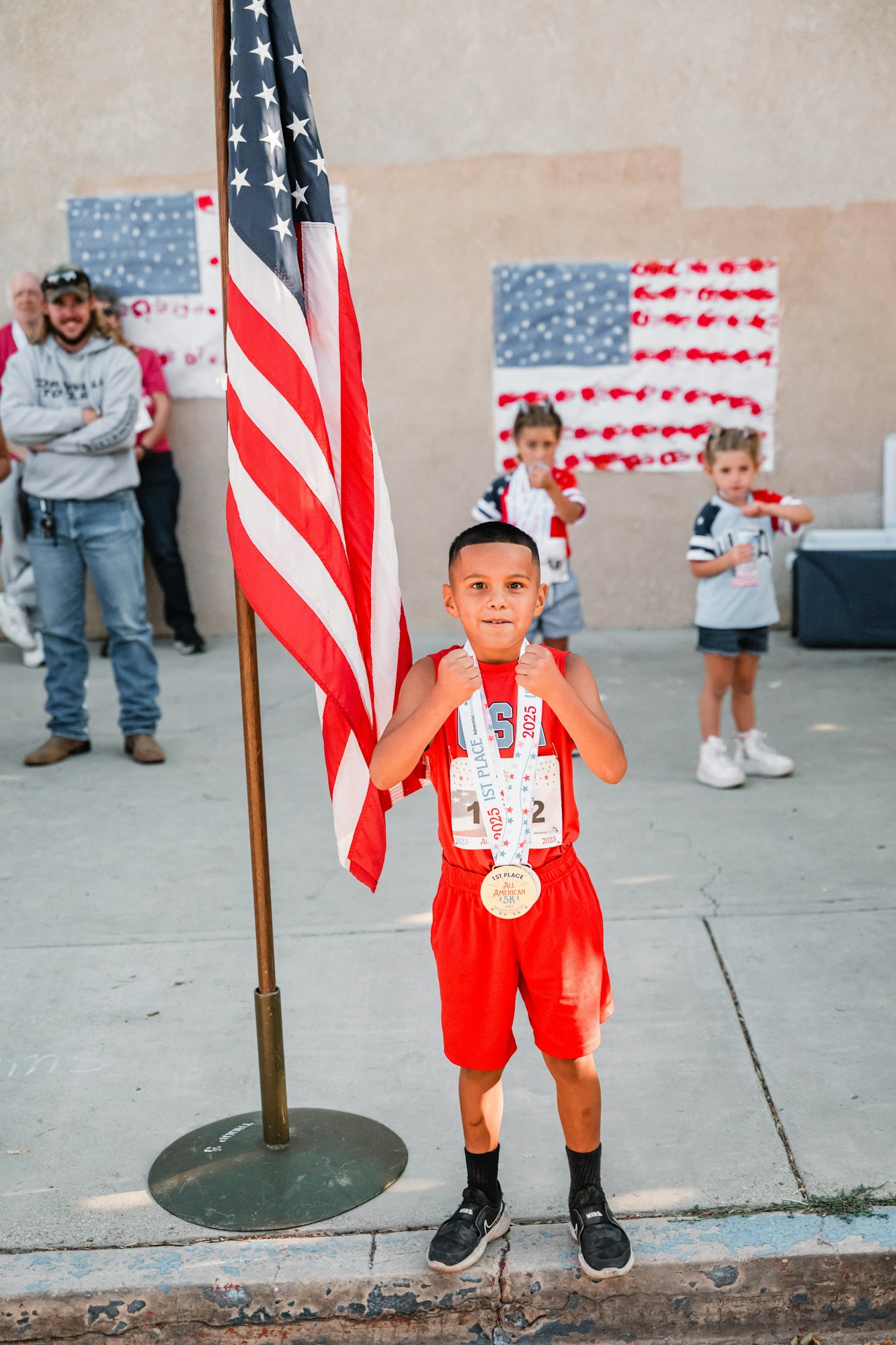 A child in red sportswear holds medals and stands by a U.S. flag, with onlookers and painted flags in the background.