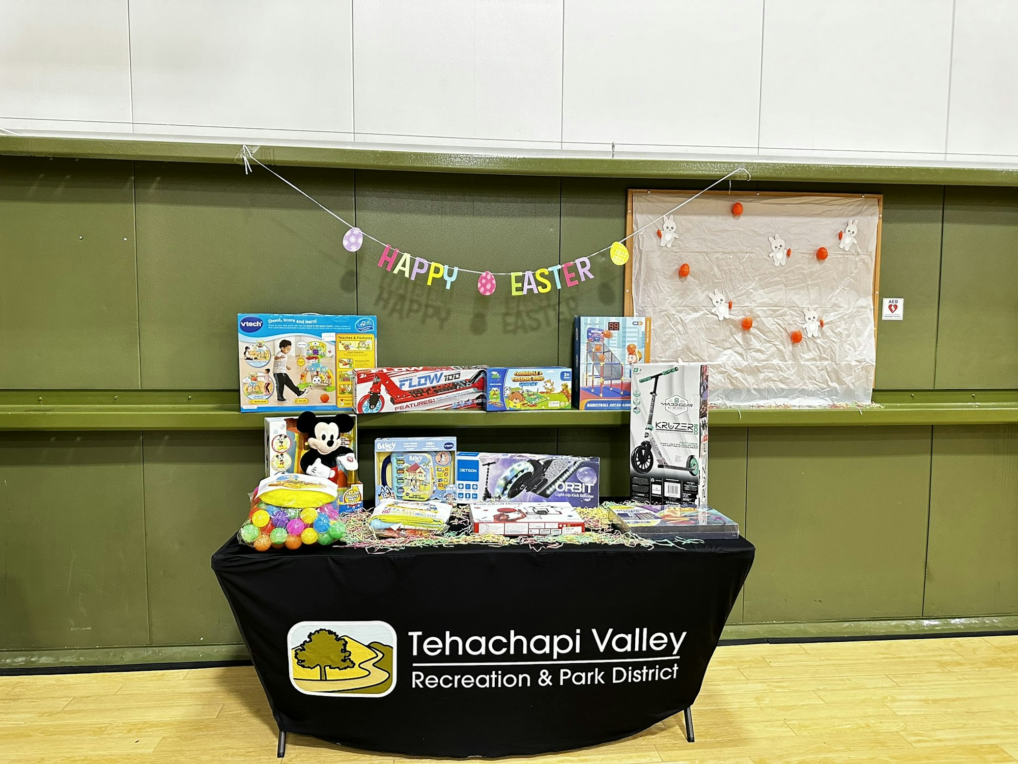 A table with toys, a Mickey Mouse plush, and Easter eggs. A "Happy Easter" banner is above, with a Tehachapi Valley Recreation logo.