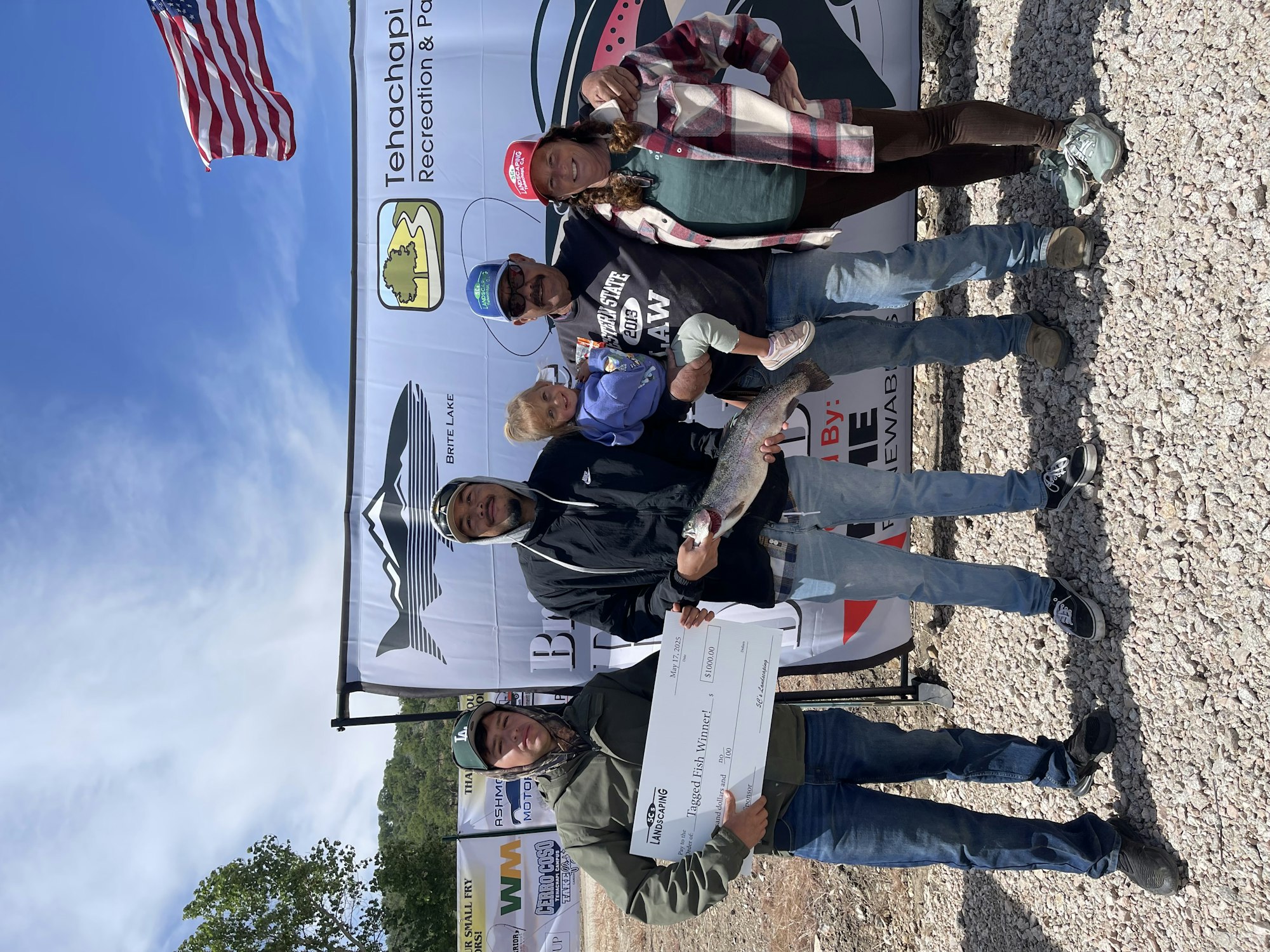 Group of people posing with a large fish and a giant check in front of a "Tehachapi Recreation" banner with an American flag.