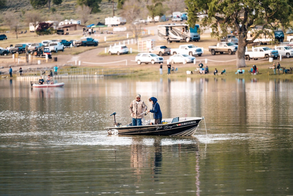 Two people in a boat fishing on a calm lake, with campers and vehicles visible on the shore.
