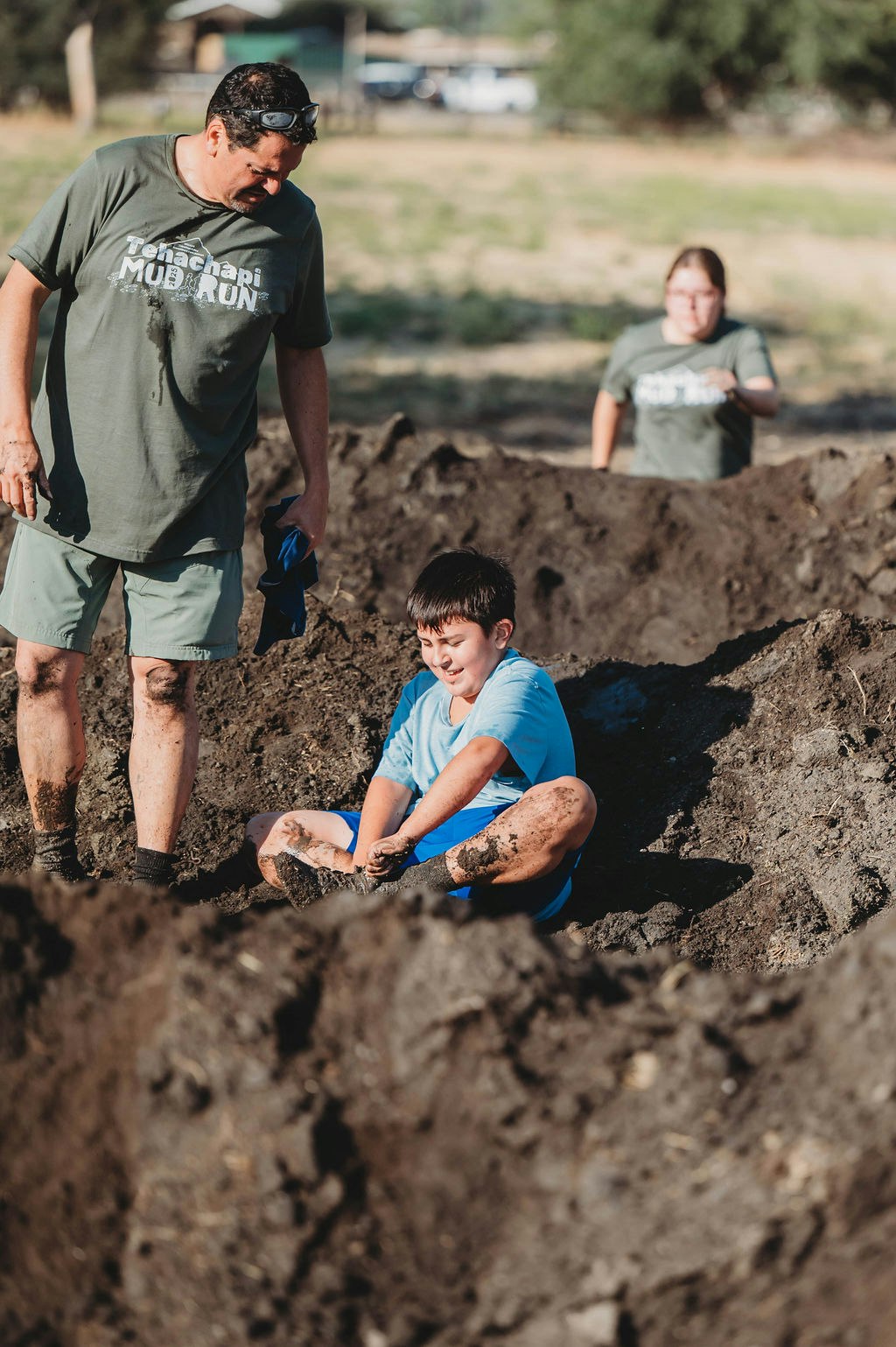 A child in a mud pit with a man standing nearby, both covered in mud, during a muddy outdoor event.