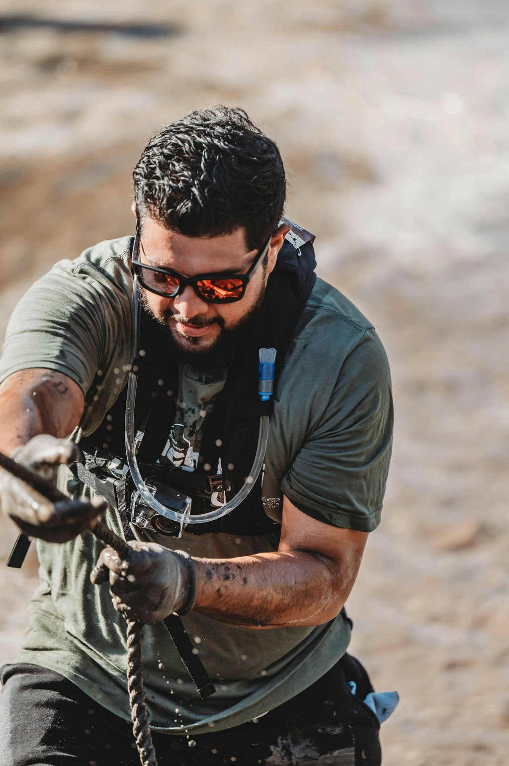 A man with sunglasses pulls a rope, wearing a hydration pack, in a muddy outdoor setting.