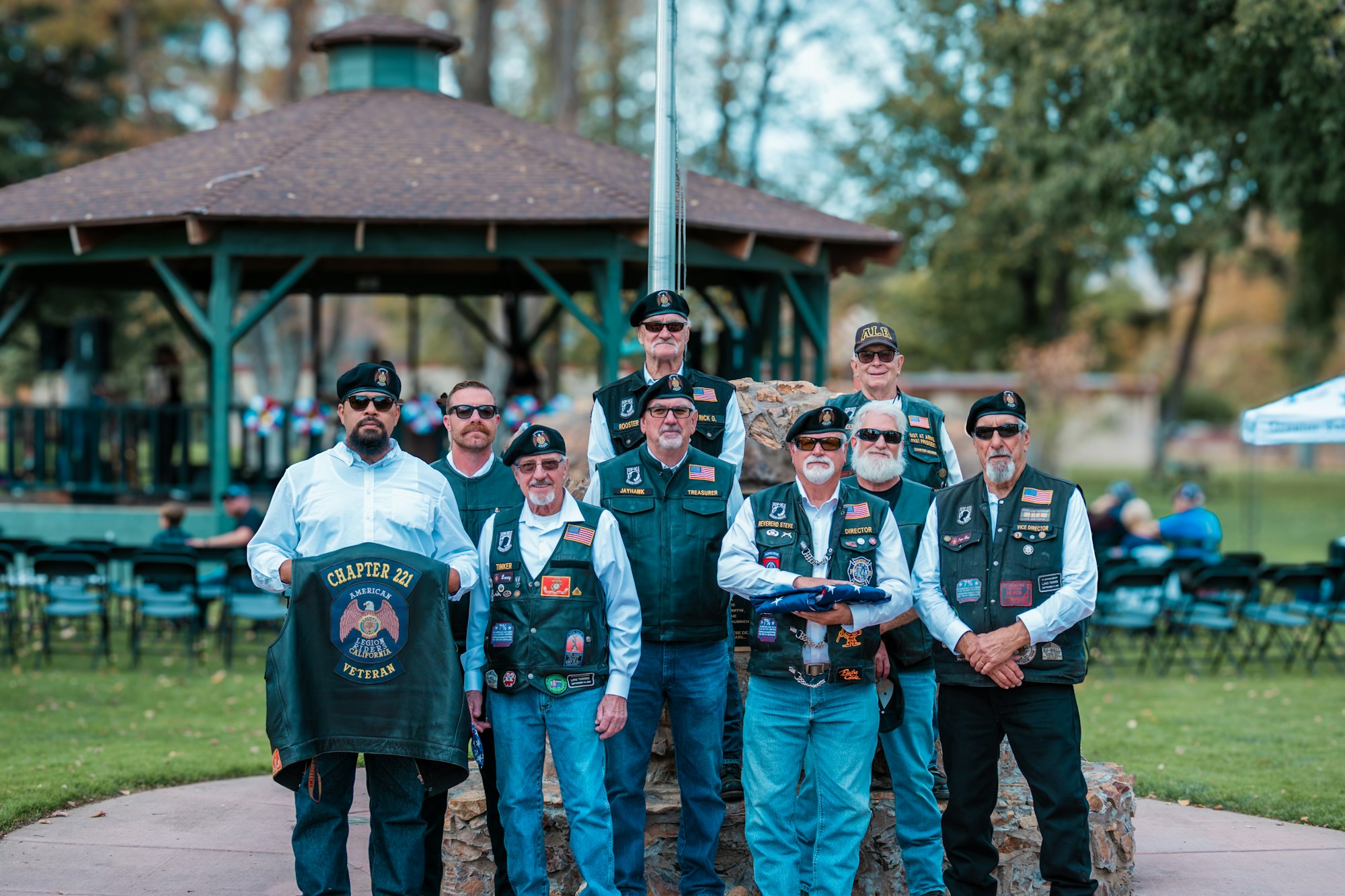 A group of older men in military-style vests and berets, standing in a park with a gazebo in the background.