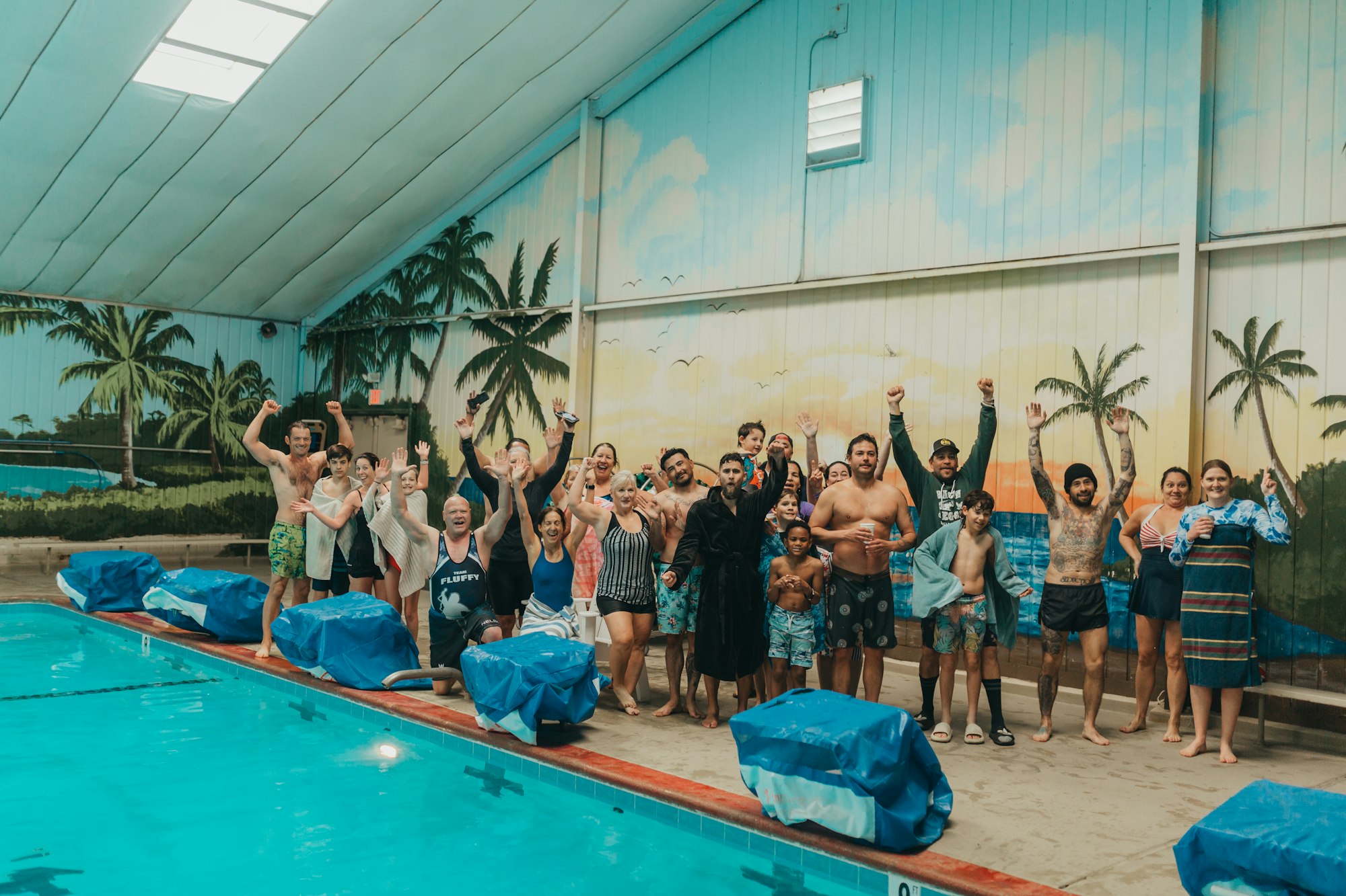 A group of people cheerfully posing by an indoor swimming pool, some with raised arms, in a celebratory mood.