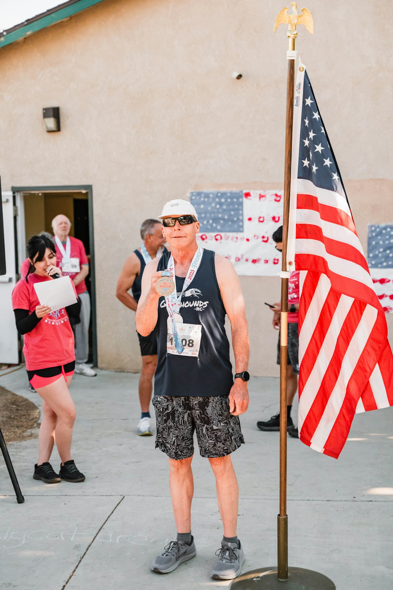 A person holding a medal stands next to a U.S. flag, wearing a race bib. Others in the background are applauding and talking.
