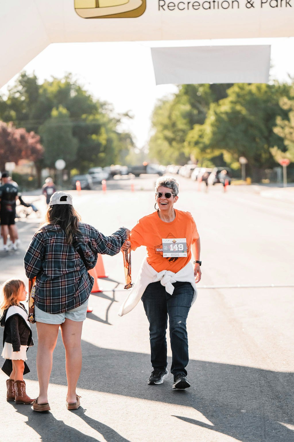 Woman in an orange shirt finishes a race, smiling and receiving a medal from another person.