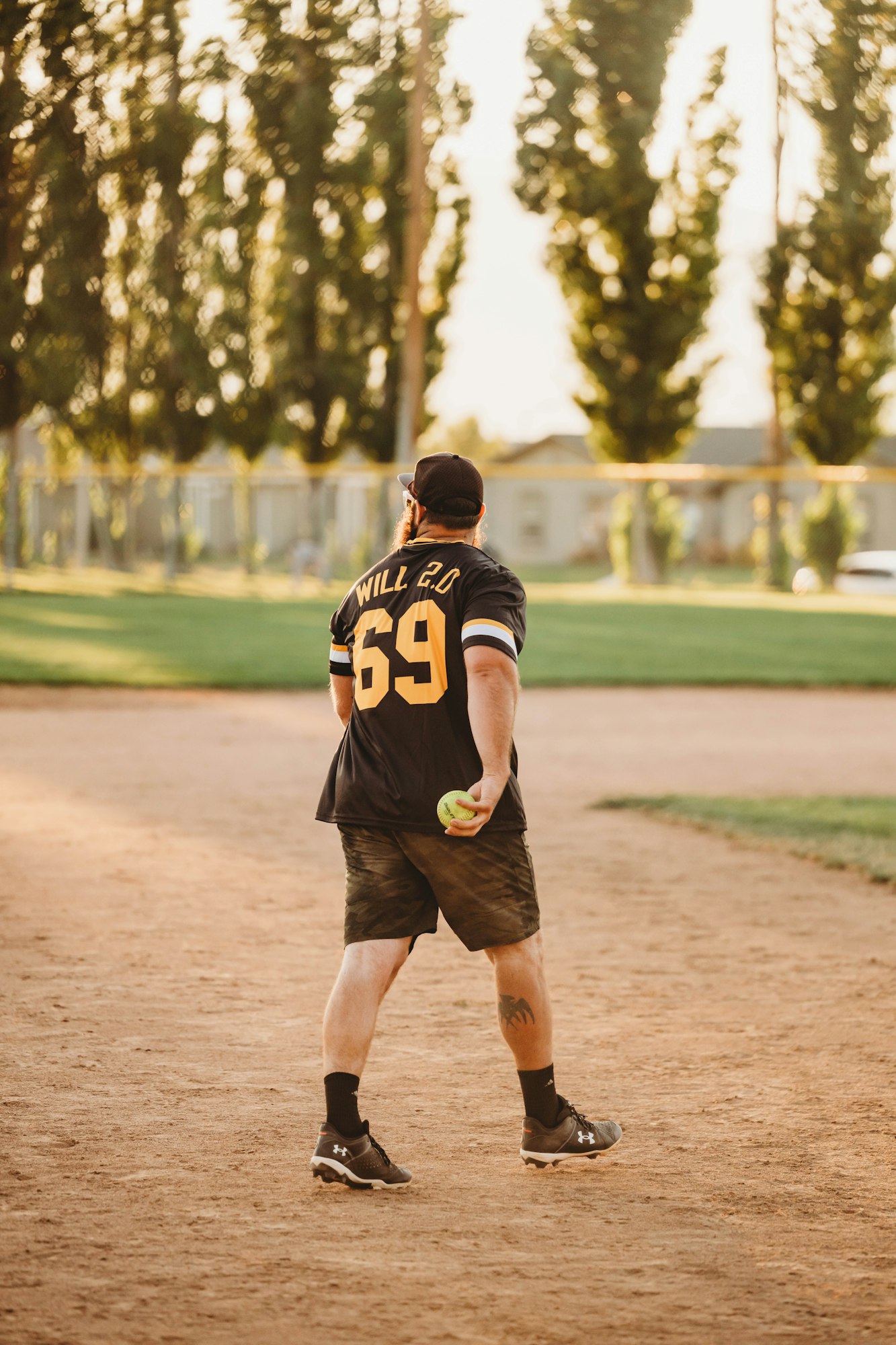 A person in a sports jersey with "69" walks on a baseball diamond, holding a softball. Trees and sunlight in the background.