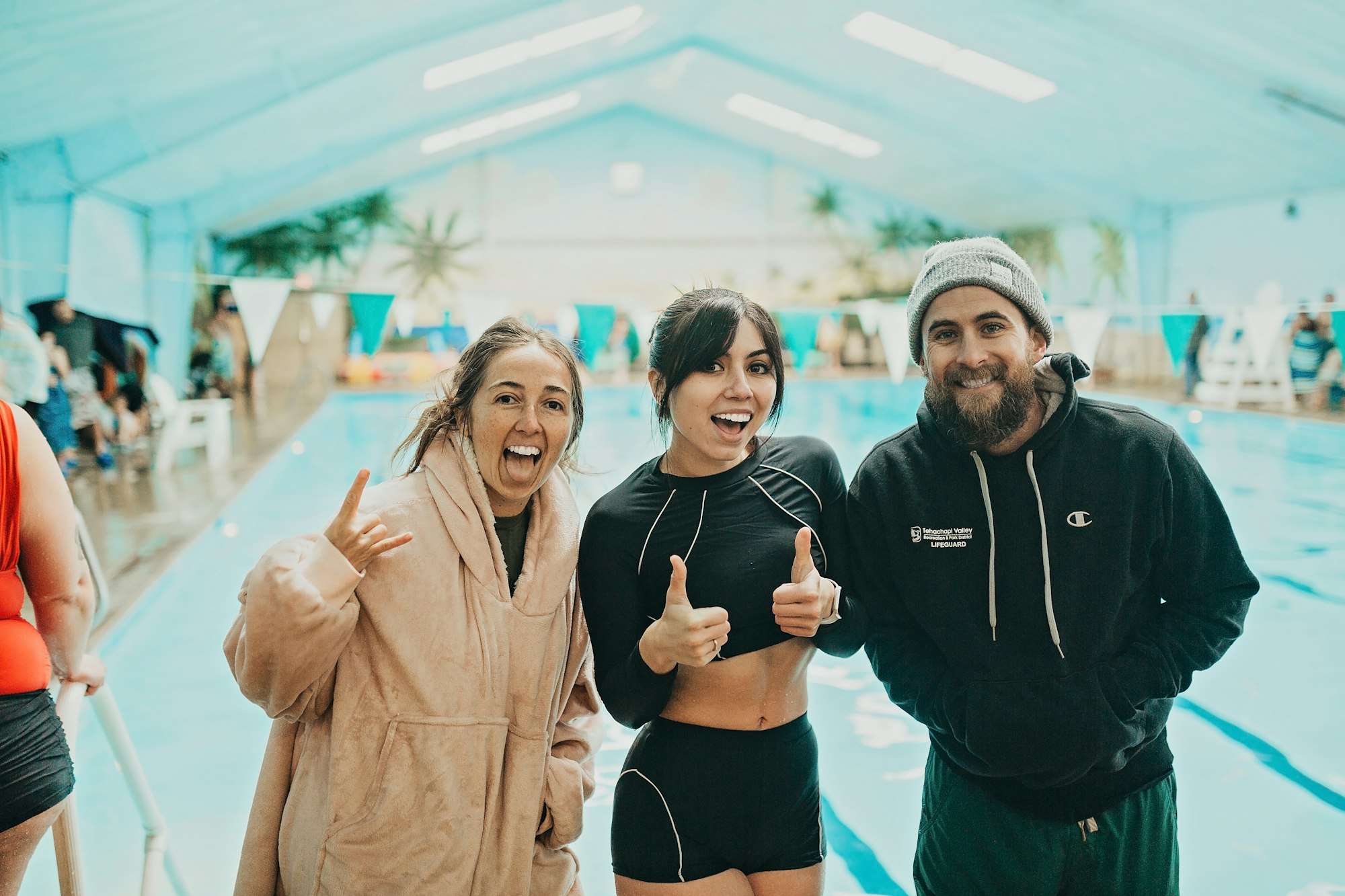 Three people smiling and posing indoors by a swimming pool.