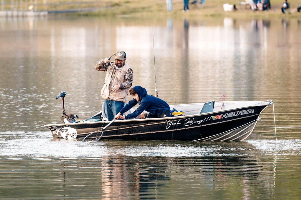 Two people on a boat, one handling a fishing rod, another with a net, on a calm lake.