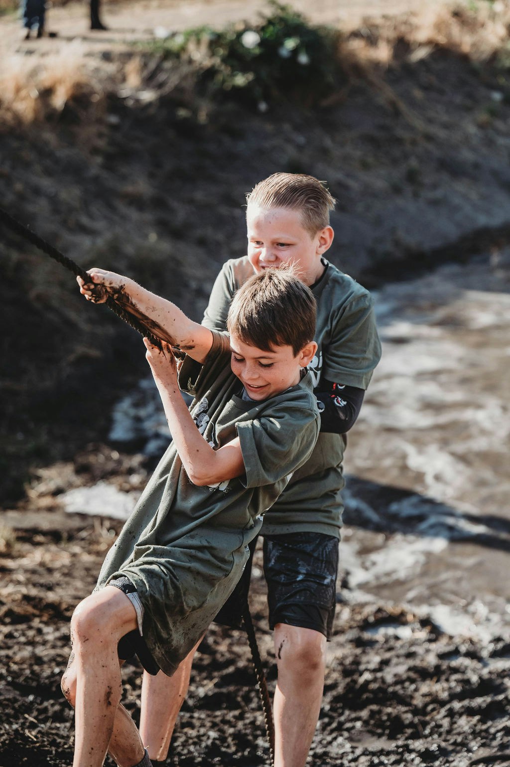 Two kids in muddy clothes pulling a rope, smiling and having fun outdoors.