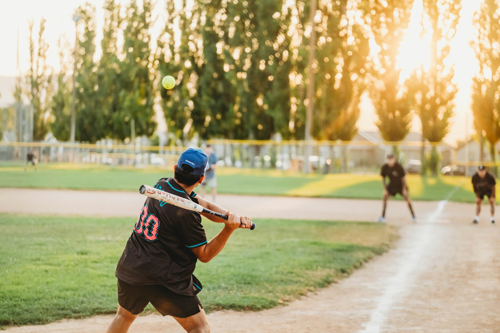 A baseball player ready to swing at a pitch, with the catcher and umpire in the background, and a sunset.