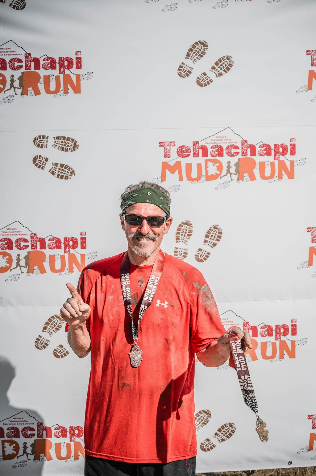 Man in a red shirt and green bandana holds a medal in front of a "Tehachapi Mud Run" backdrop, smiling and gesturing with one finger.