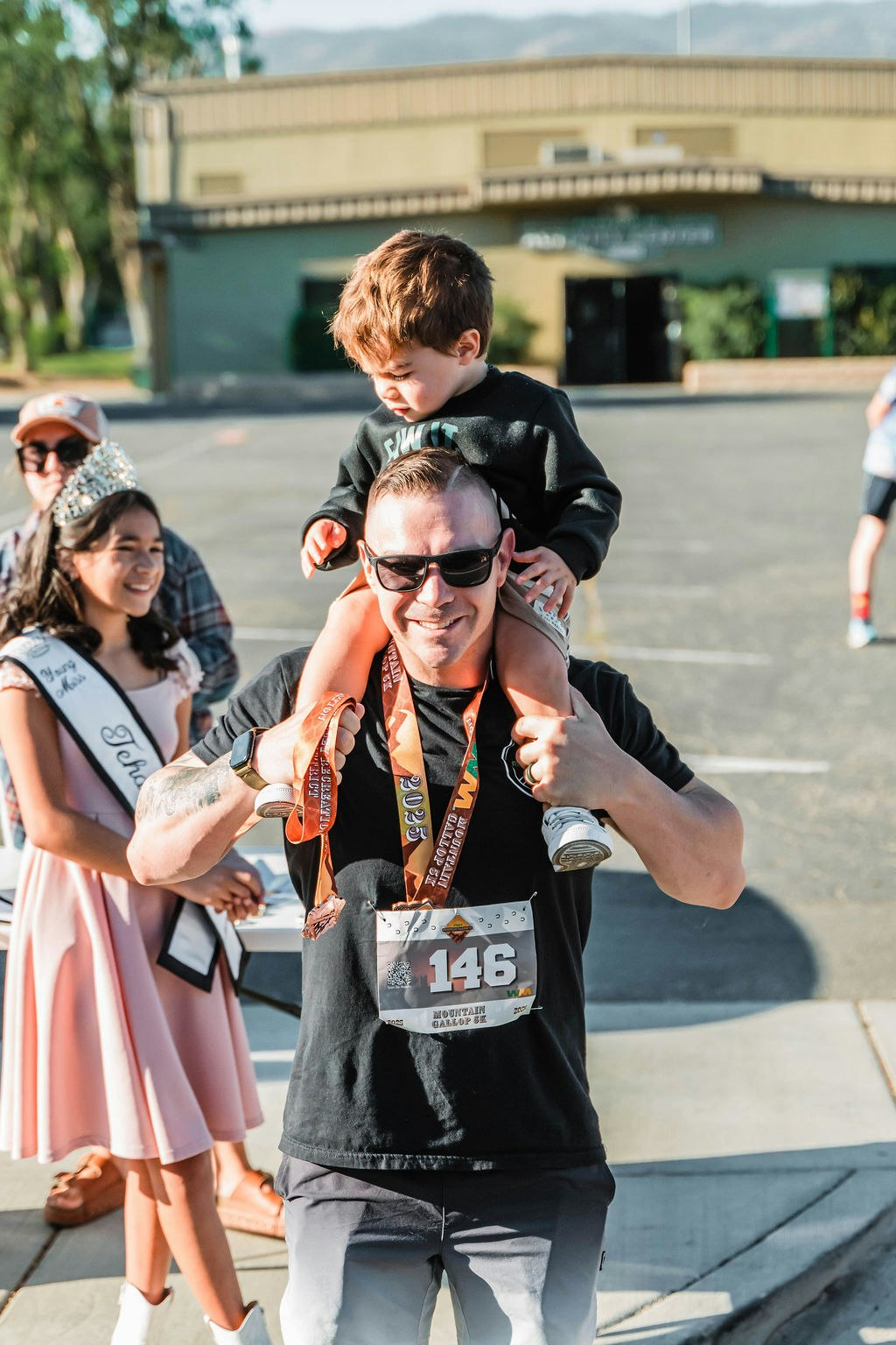 A man with sunglasses carrying a child on his shoulders, wearing race medals. People in the background, one with a sash and crown.