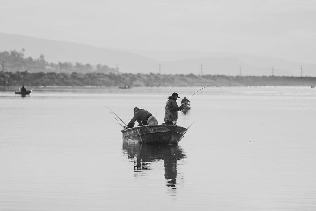 Two people fishing from a boat on calm water in a black and white photo.