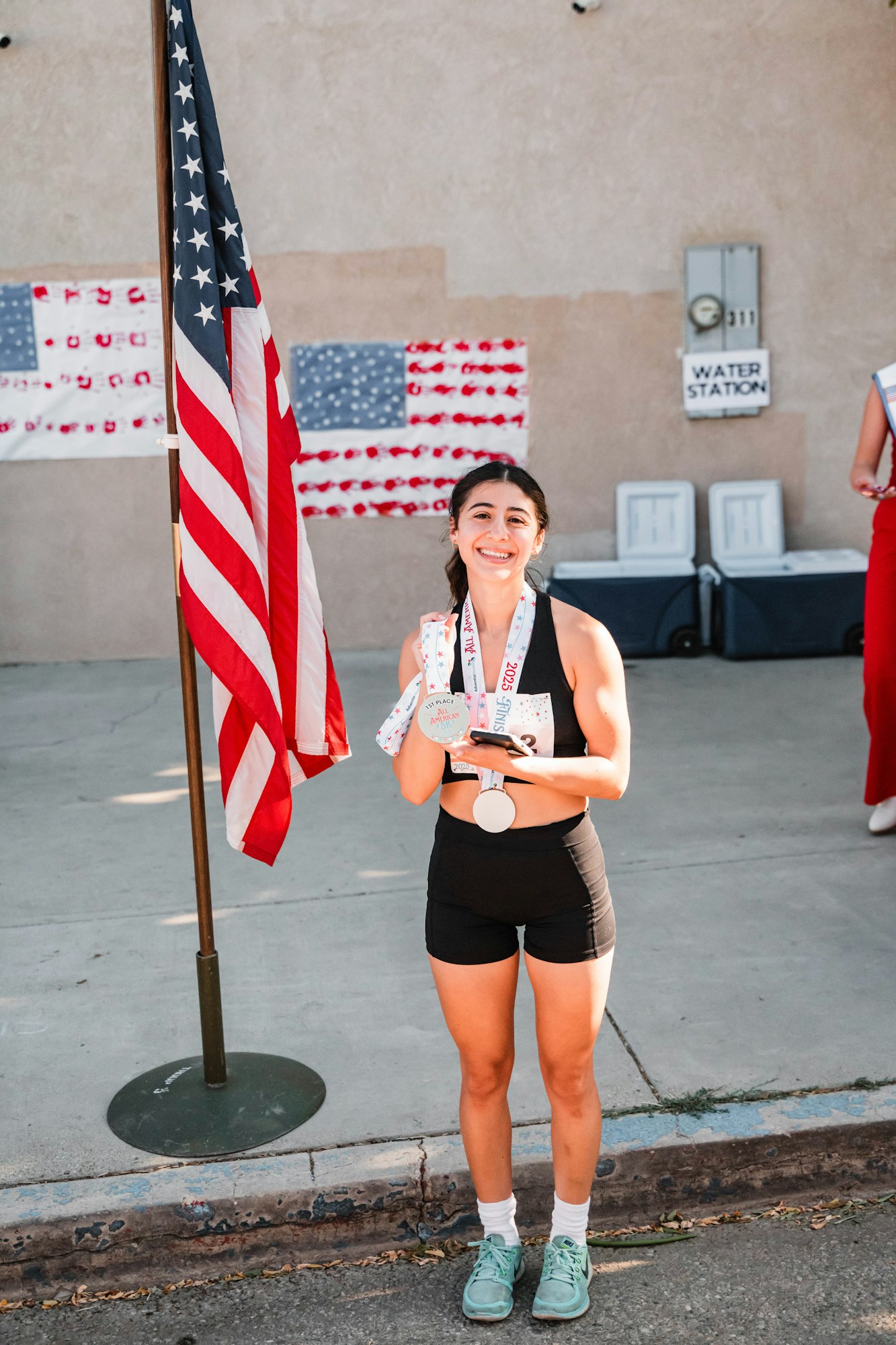 Person smiling with medals, standing by a U.S. flag and a water station sign.