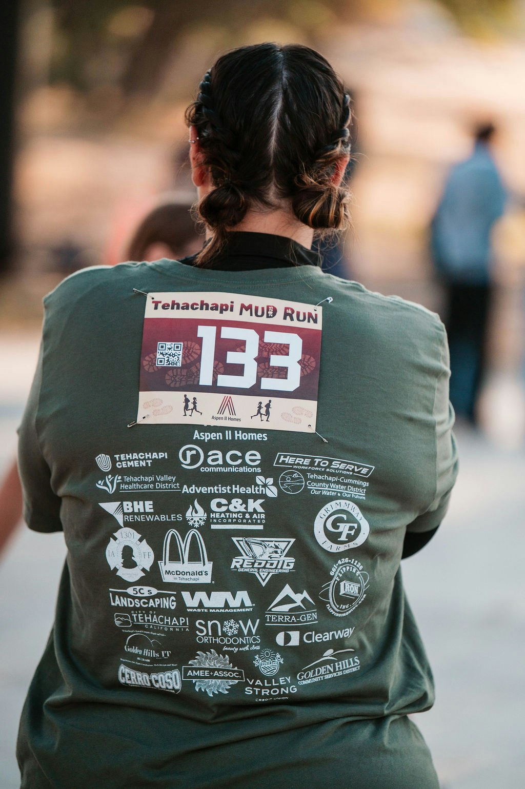 Person with braided hair wearing a green shirt and "Tehachapi Mud Run" bib number 133, with sponsor logos on the back.