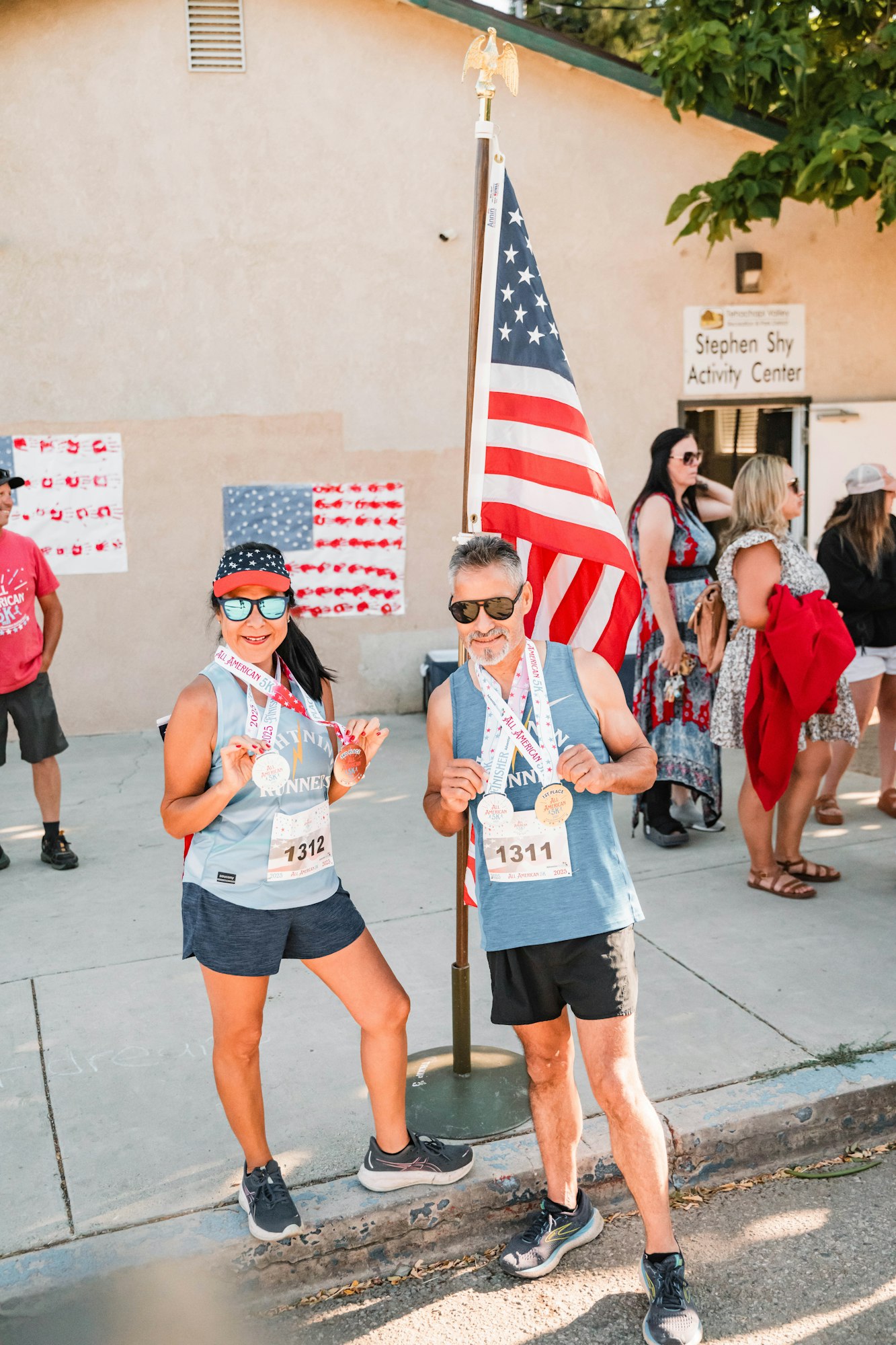 Two people with medals, race bibs 1311 and 1312, posing by a U.S. flag. Others and activity center in background.