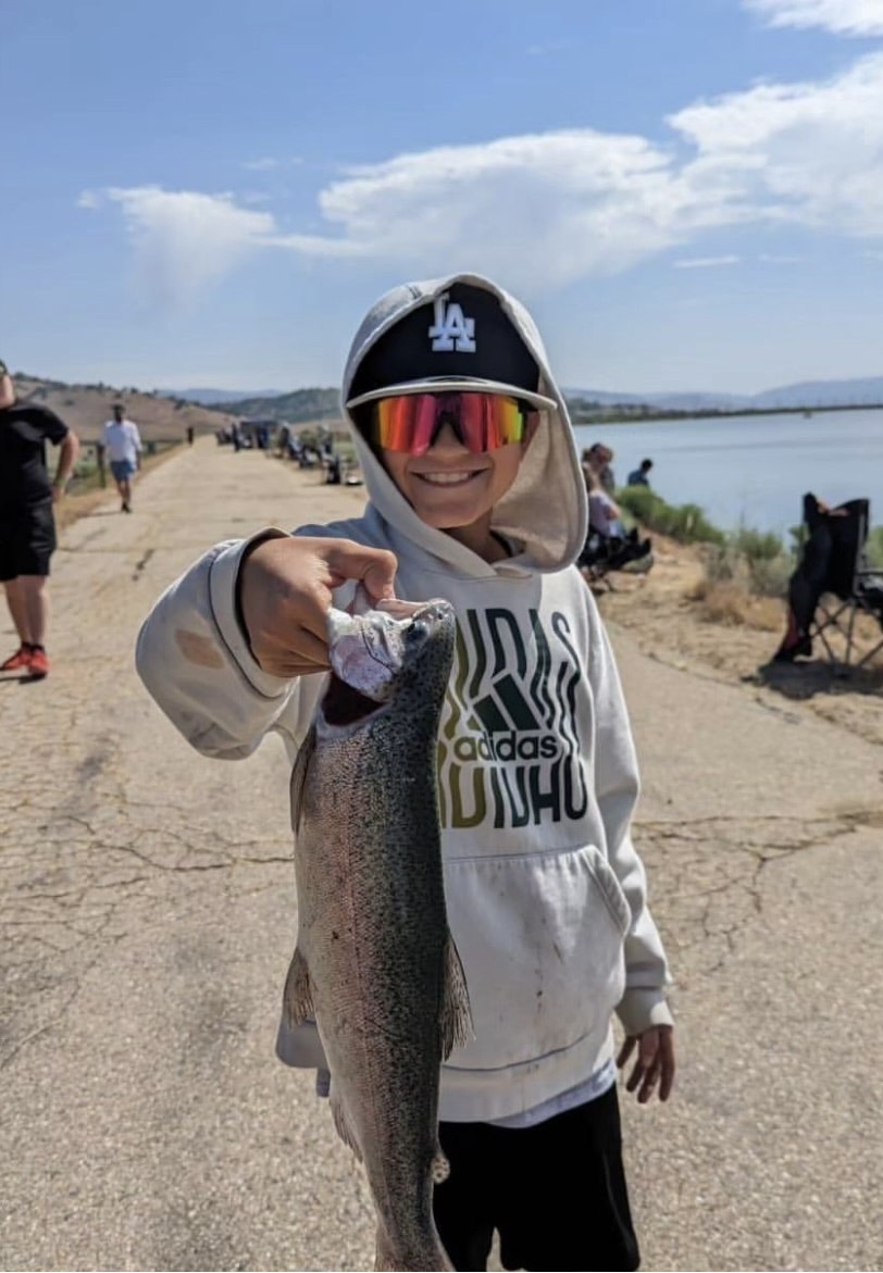 A person wearing sunglasses and a hat holds up a fish, with a lake and people in the background.