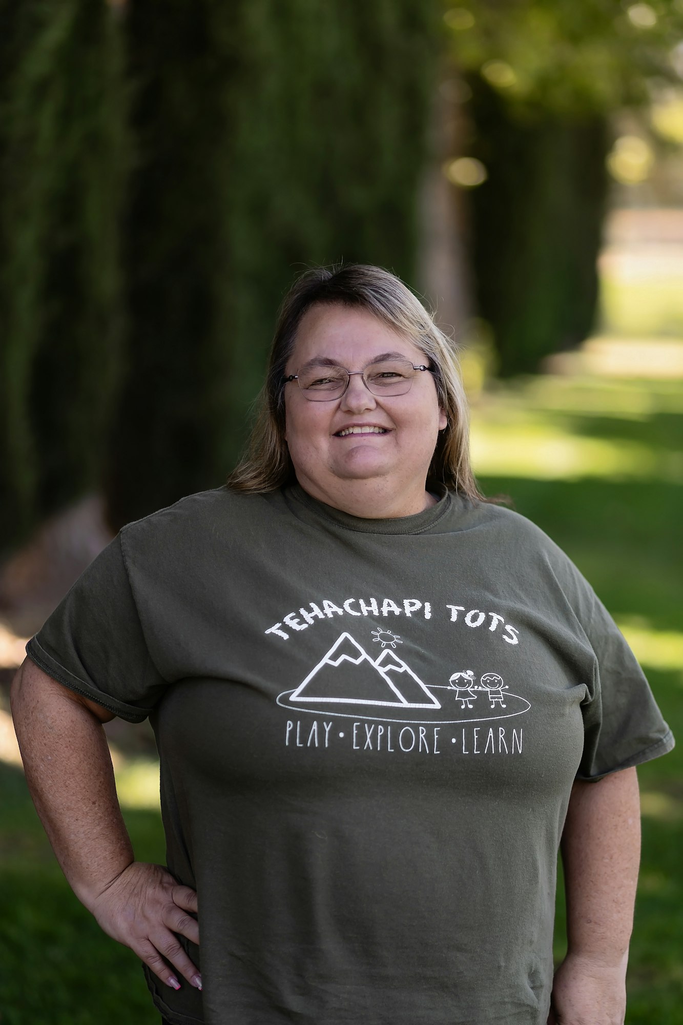 A person smiling outdoors in a "Tehachapi Tots" T-shirt with play, explore, learn graphics.