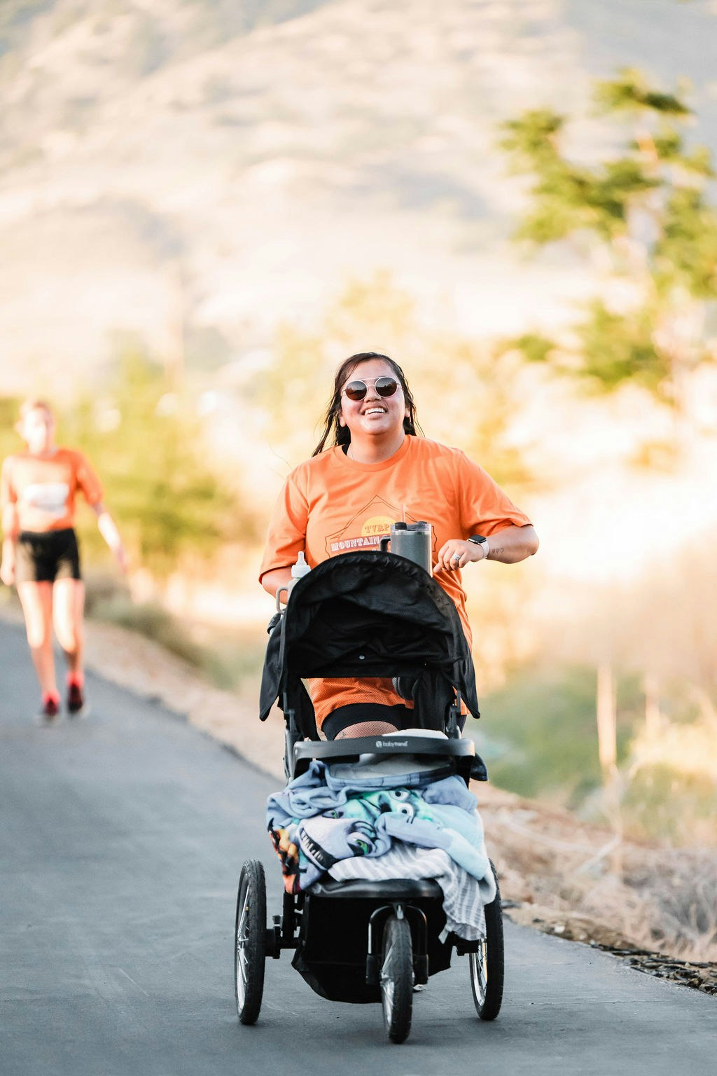 Person smiling, pushing a stroller on a sunny pathway, wearing an orange shirt and sunglasses.