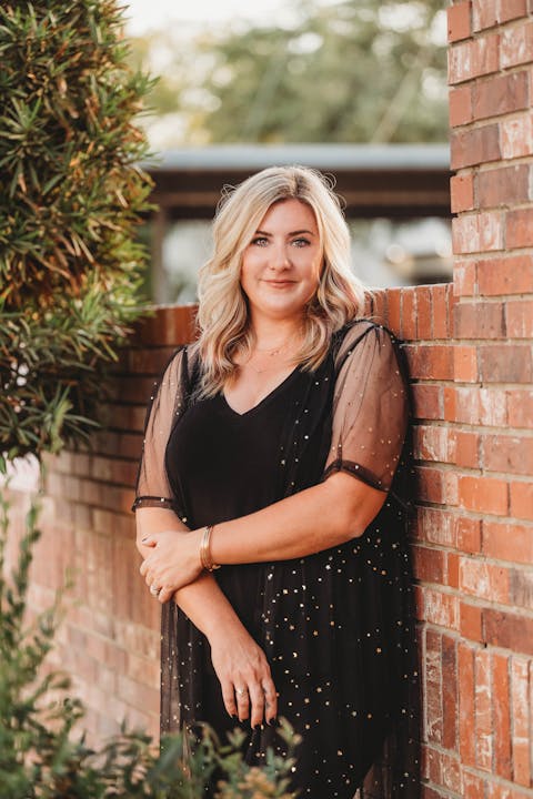 A woman in a black dress with gold accents stands beside a brick wall, greenery to the left.