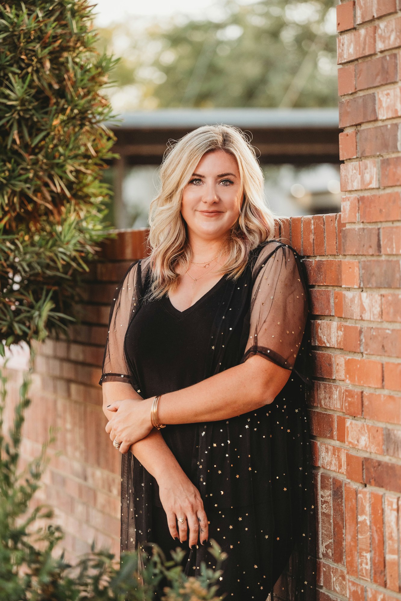 A woman in a black dress with gold accents stands beside a brick wall, greenery to the left.