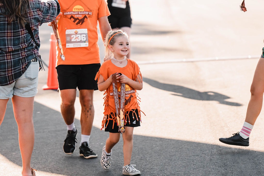 A young child in an orange T-shirt holds a medal, smiling while walking with others during an outdoor event.