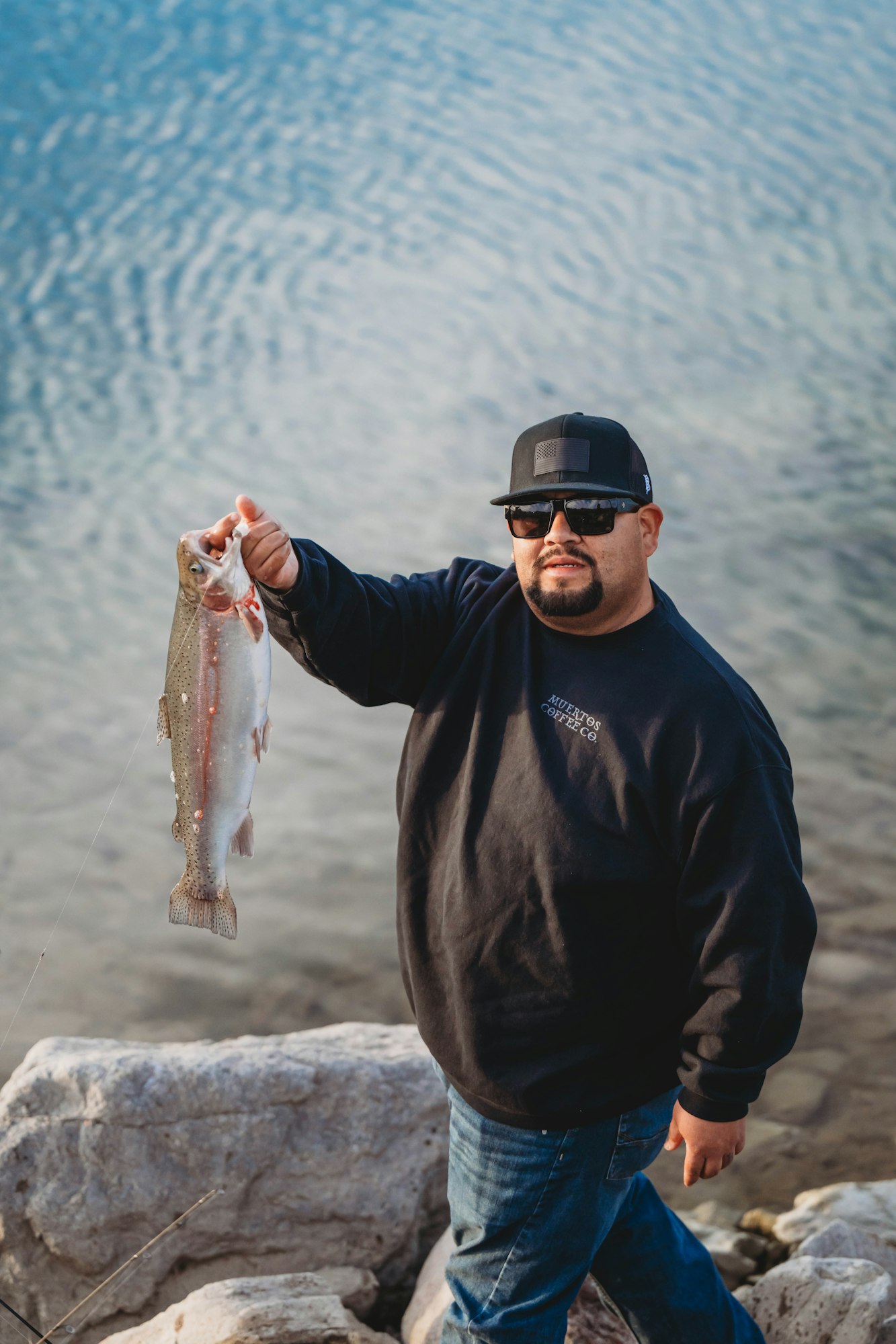 A person in sunglasses and a cap holding a fish by a lake.