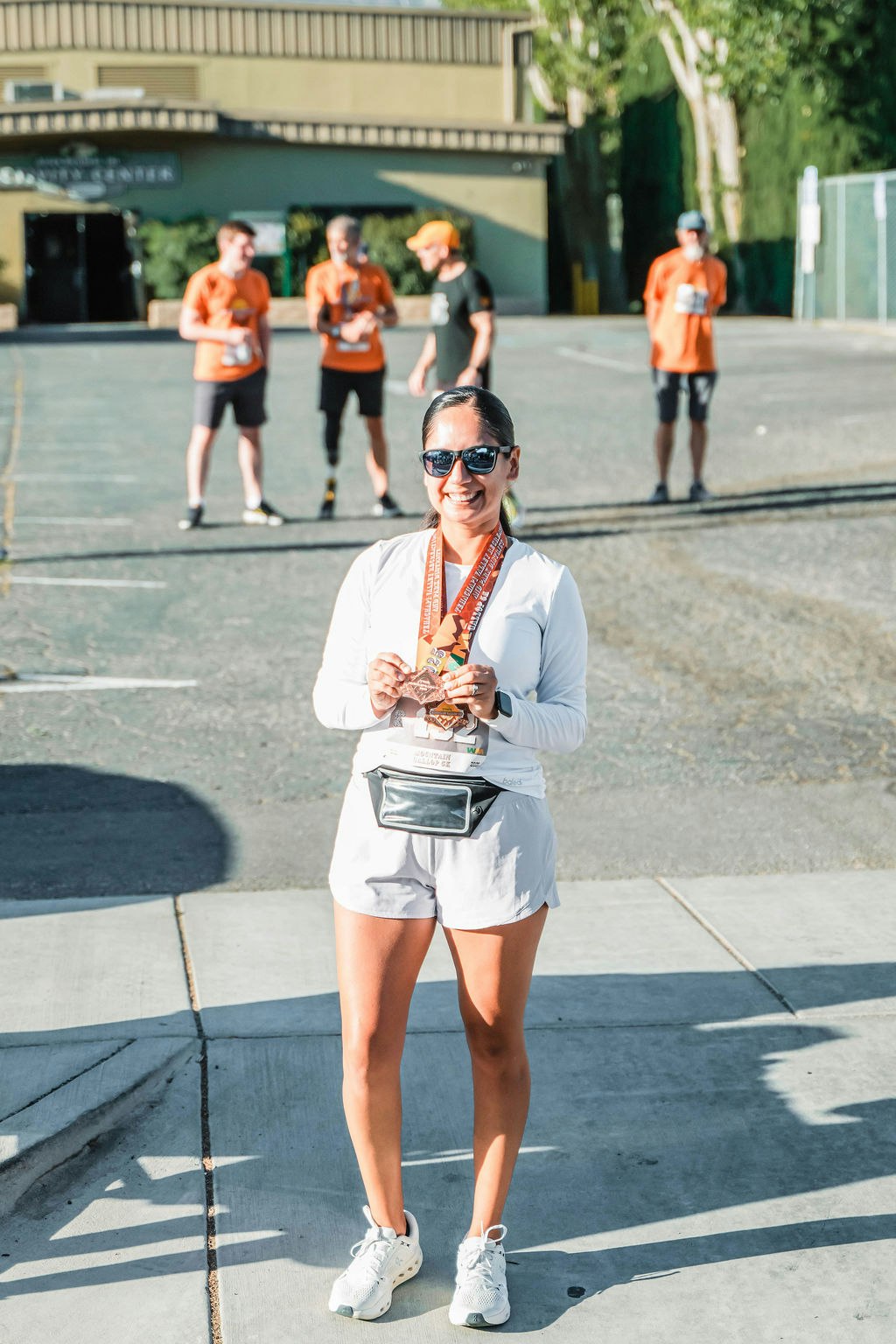 A smiling woman with medals stands in front of a building, with three people in orange shirts in the background.