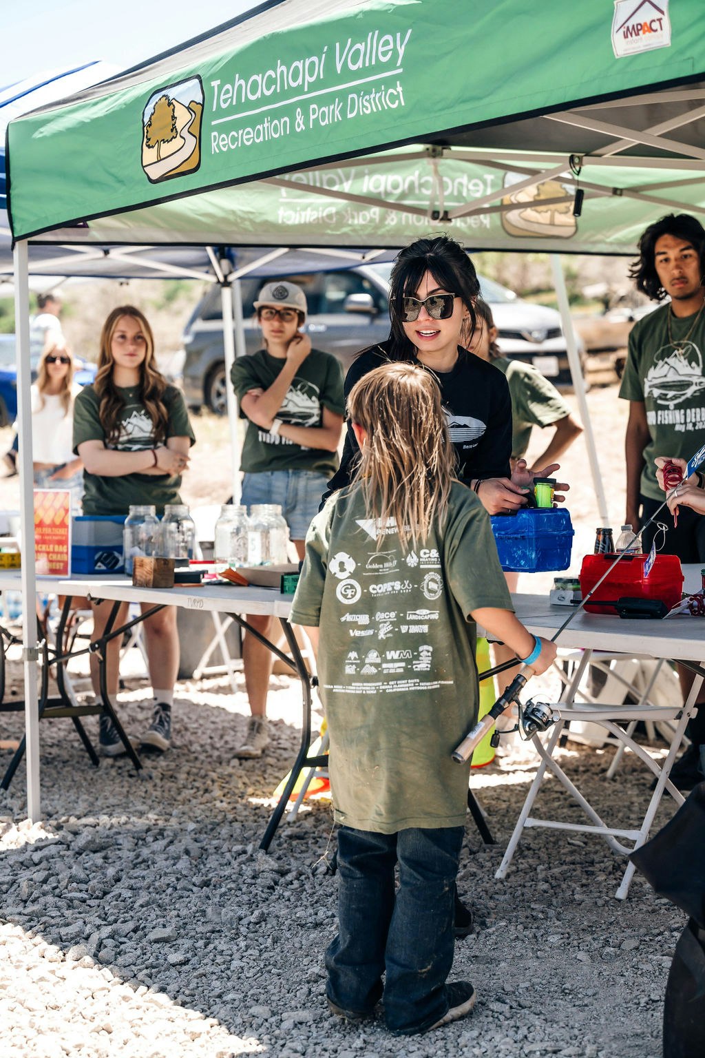 People at an outdoor event under a tent; a child with a fishing rod is in the foreground.