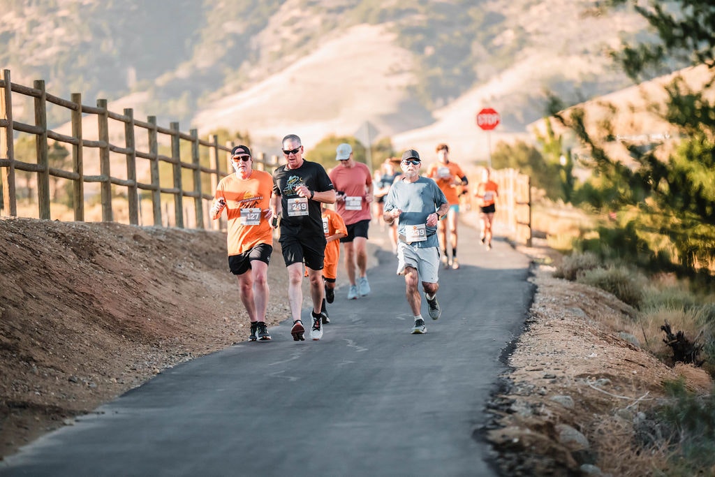 People running on a paved path during a race on a sunny day, with hills and trees in the background.