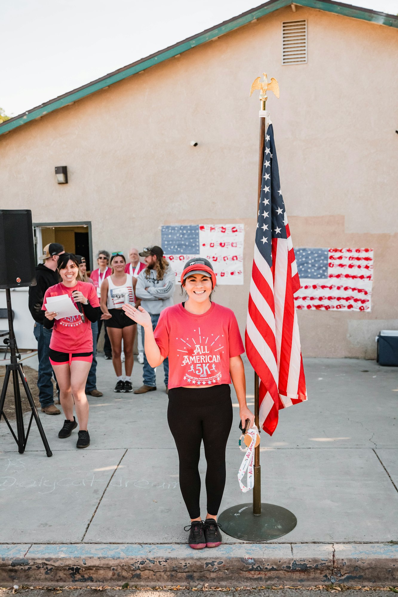 A smiling woman stands beside an American flag, wearing a “5K” shirt, holding a medal. People in the background with American-themed decor.