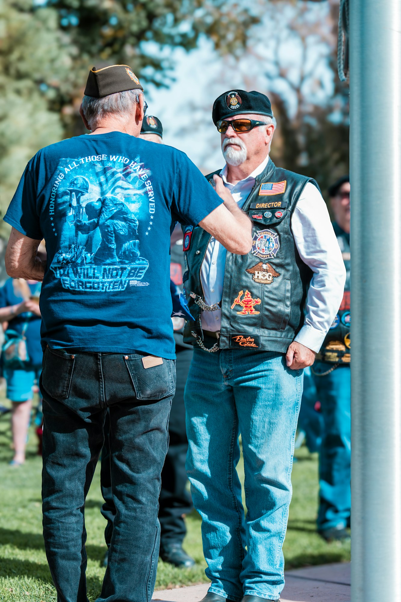 Two men in military-themed attire conversing outdoors, one wearing a vest with patches and another in a blue t-shirt honoring veterans.
