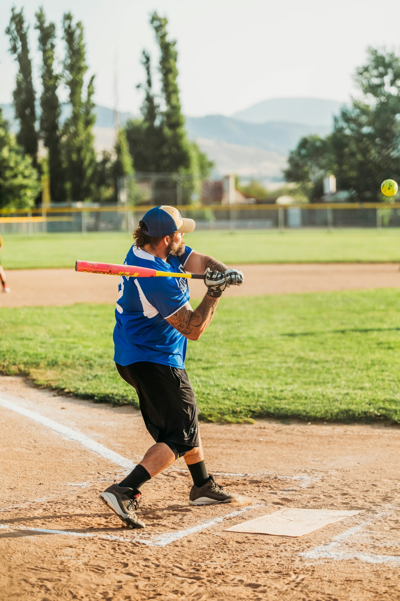 A person playing softball, swinging at an incoming ball, in a sunny, outdoor setting with trees in the background.