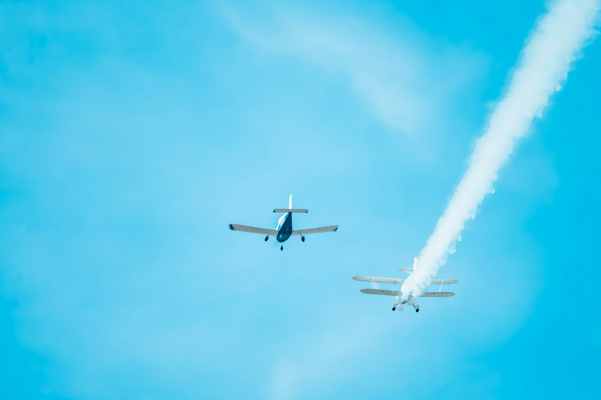 Two small airplanes flying in blue sky, one with a smoke trail.