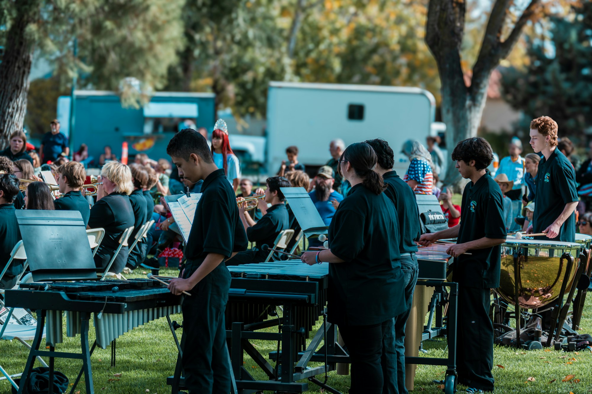 Outdoor band performance with musicians playing percussion instruments in a park, surrounded by an audience.