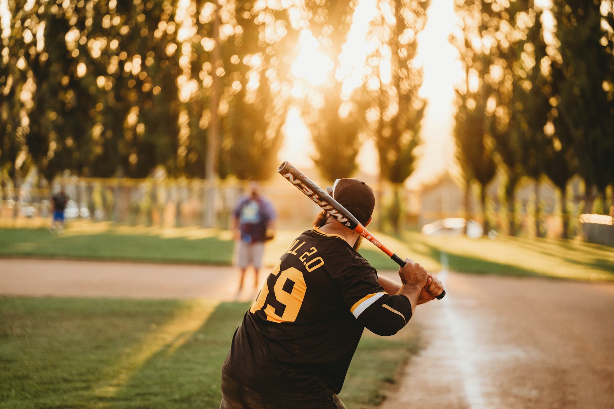 A baseball player ready to swing at sunset, with trees and a person in the background.