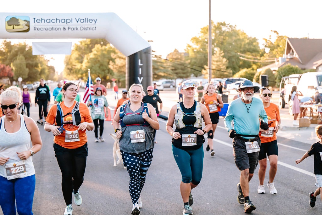 A group of people running a race under a "Tehachapi Valley Recreation & Park District" arch.