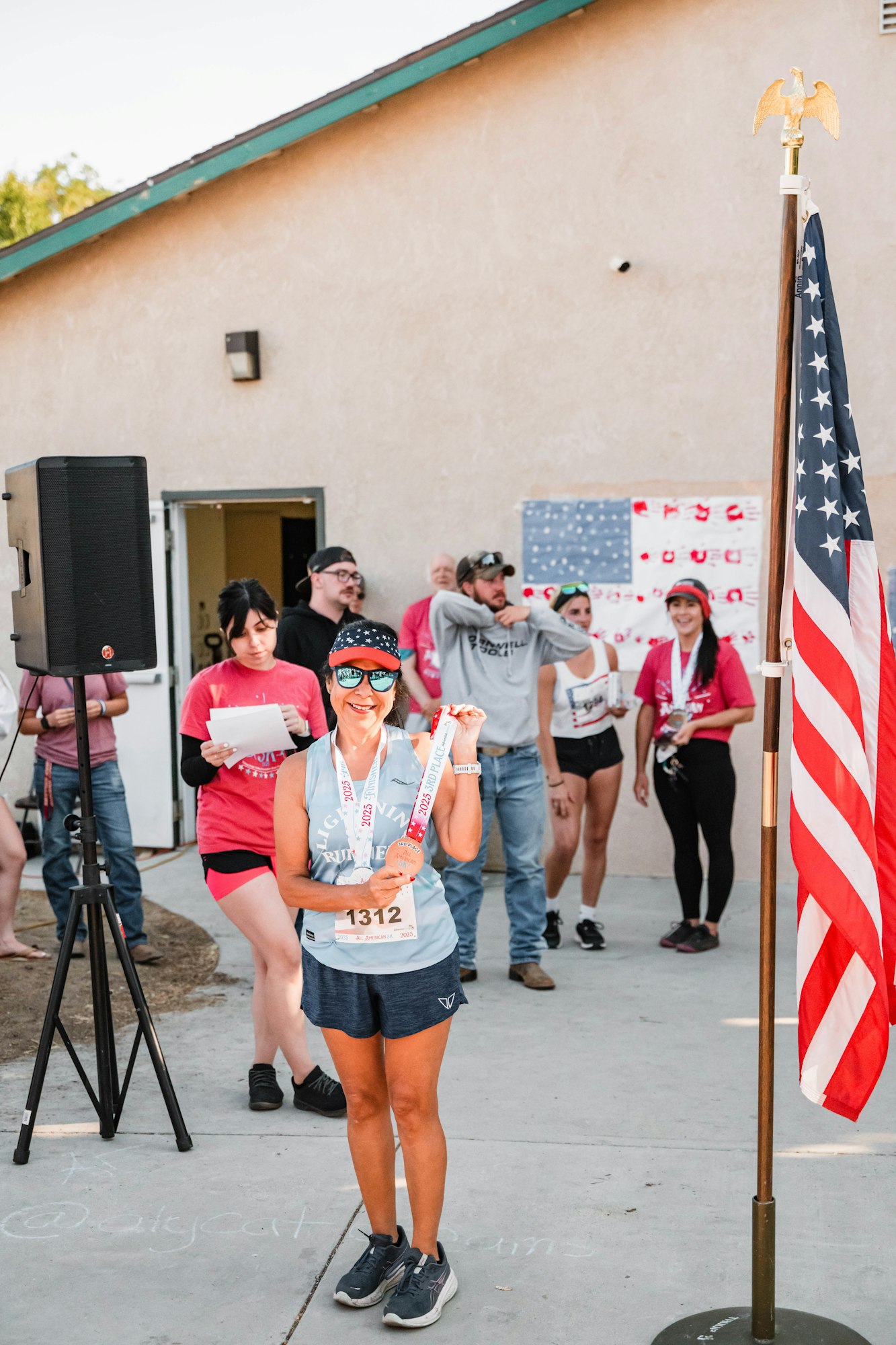 A woman with race gear and medals poses, people and a flagpole with the U.S. flag behind her.