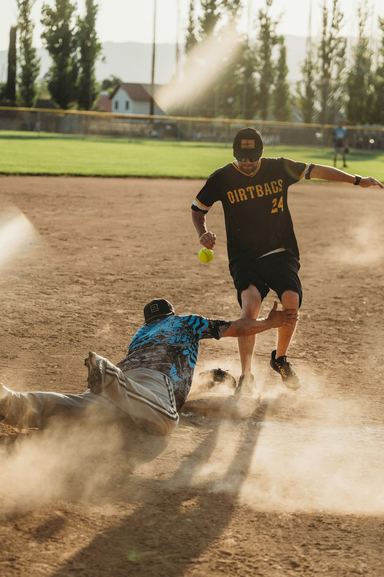 A softball player sliding into base while another tries to tag him out, amidst dust and sun glare.