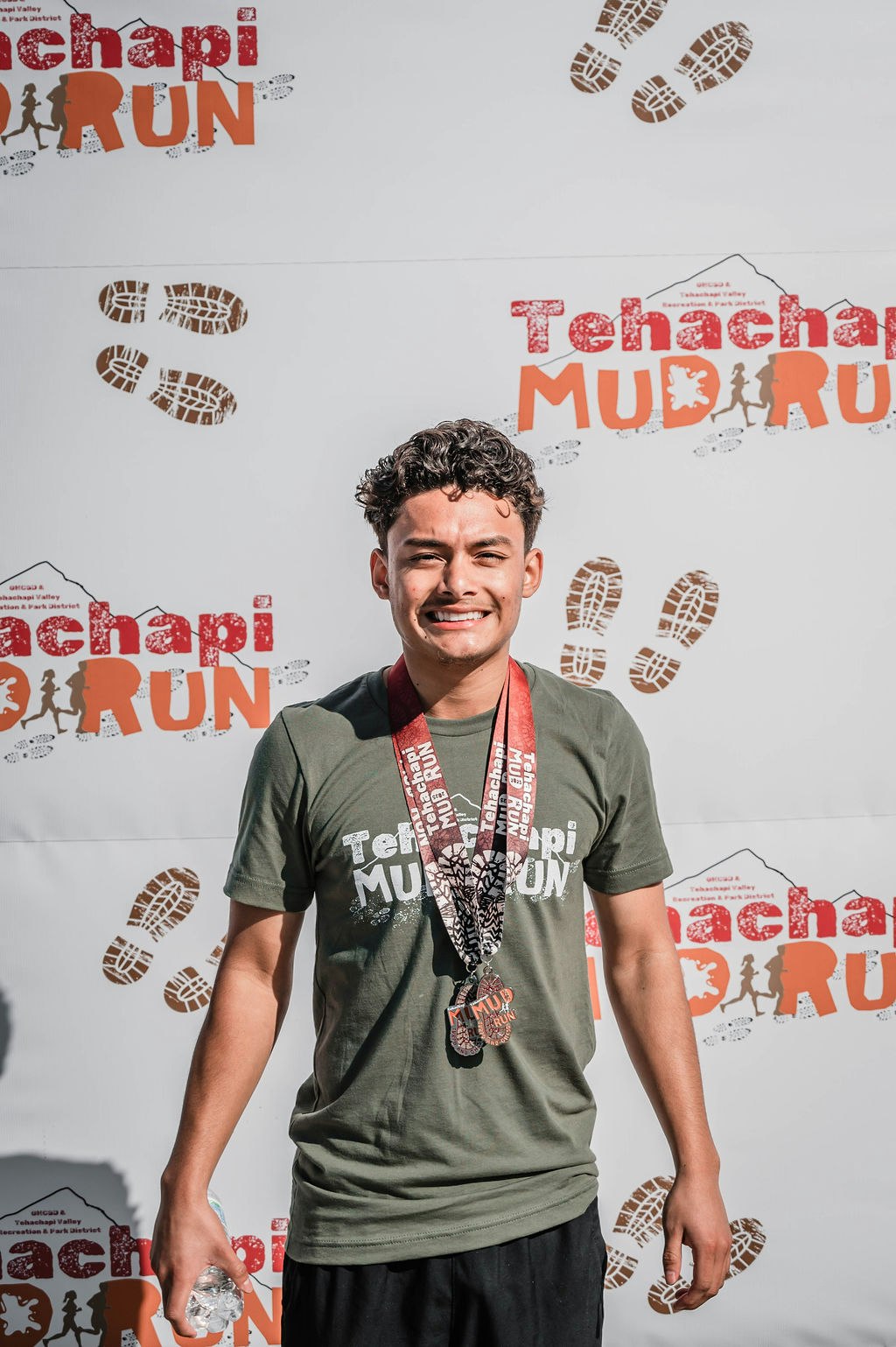A person smiling with a medal, wearing a "Tehachapi Mud Run" shirt, standing against a backdrop with the same event logo.