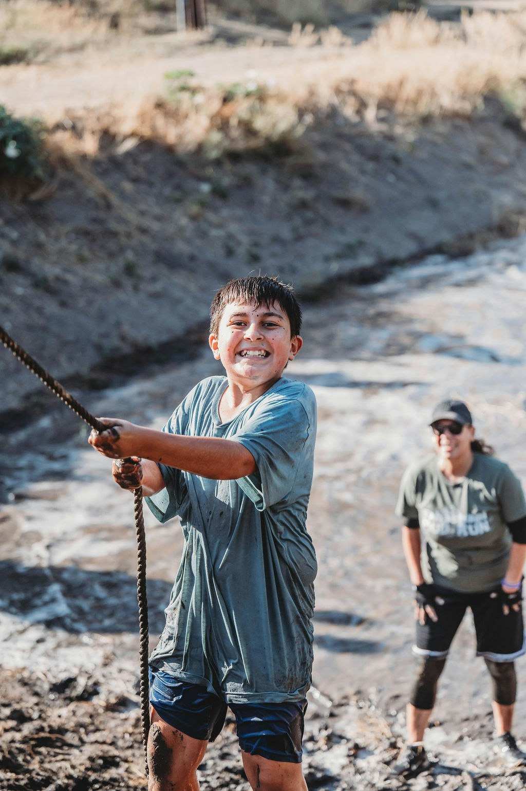 A child is smiling while climbing a muddy hill with a rope, another person watches in the background.
